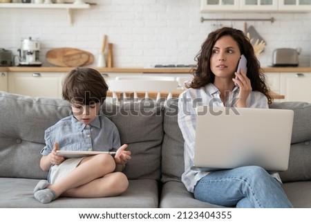 Busy woman mom with kid work from home during covid-19 quarantine lockdown. Young businesswoman talk on mobile phone, use laptop while little son play on digital tablet. Career and motherhood concept