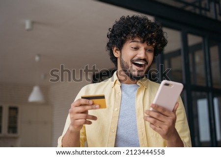 Amazed Indian man holding credit card using mobile phone shopping online. Young happy freelancer receive payment, check card balance. Emotional guy ordering food online looking at digital screen