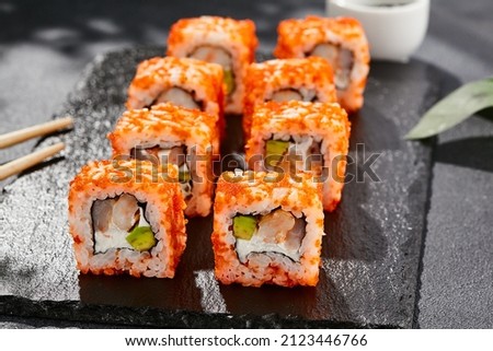 Maki sushi on dark stone table. California maki with tobiko. Sushi roll with cheese,shrimp, avocado inside, maguro outside. Style concept japanese menu with black background, leaves and hard shadow