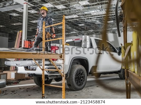 Caucasian Commercial Building Construction Worker in His 30s Wearing Hard Hat and Keeping Tools in His Hands Staying on His Pickup Truck Cargo Bed Royalty-Free Stock Photo #2123458874