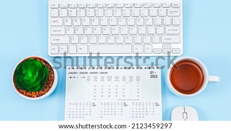 Calendar for January 2022 with a keyboard, mouse, a mug of tea and a small pot with a flower on a light blue background, close-up top view.