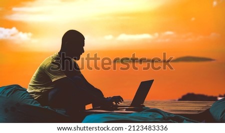 A silhouette portrait of a man working on his laptop with the background of the sea the sky and an island behind him during sunset Royalty-Free Stock Photo #2123483336