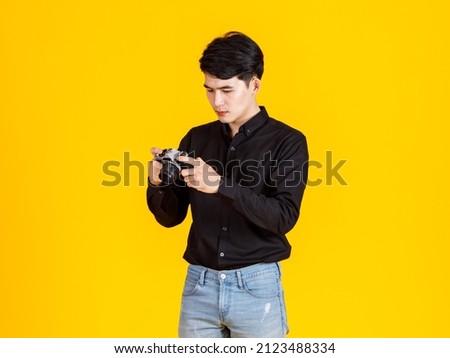 Portrait studio shot of millennial Asian young professional male fashion photographer journalist in casual outfit standing smiling holding aiming digital DSLR mirrorless camera on yellow background.