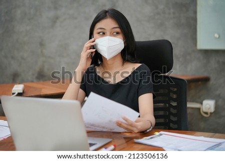 An Asian business woman was calling a client while looking outside. She works and wears a mask, holding her documents.                             Royalty-Free Stock Photo #2123506136