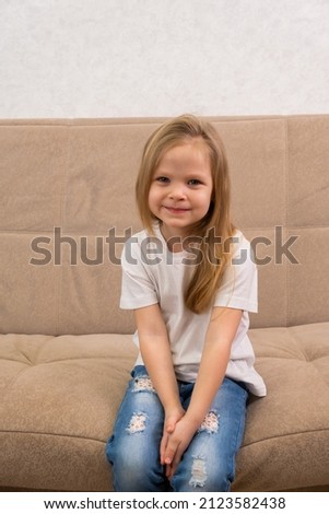 Portrait of a little blonde girl in a white T-shirt. Sitting on the couch and smiling.