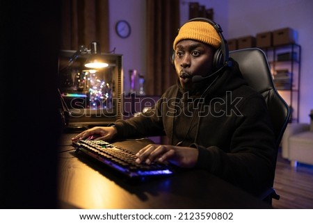 A shocked man is playing an emotional computer game, reliving the rounds, clicking fingers on the backlit keyboard, his eyes staring at the monitor, the room lit by LEDs