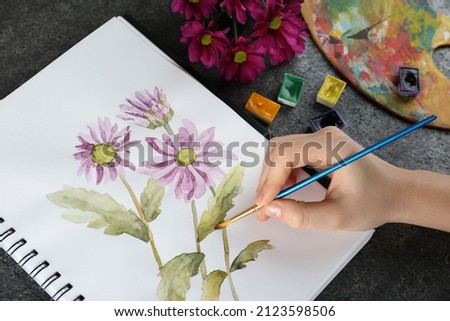Woman drawing beautiful chrysanthemum flowers in sketchbook at grey table, closeup