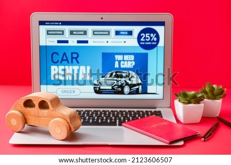 Laptop with open page of car rental site, passport and wooden toy on red background
