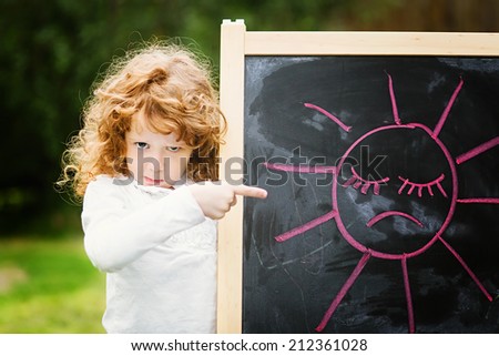 The sad girl points a finger at the picture with an unhappy sun, painted in red chalk on a blackboard. Educational concept. 