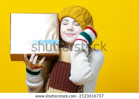 Dreaming Smiling Caucasian Teenage Girl in Warm Knitted Hat and Warm Scarf Holding Wrapped Golden Giftbox and Looking Upwards Over Yellow Background.Horizontal Image Composition