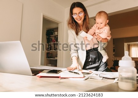 Cheerful mom holding her baby while working on a new project in her home office. Happy single mom smiling at the swatches on her desk. Female interior designer balancing work and motherhood.