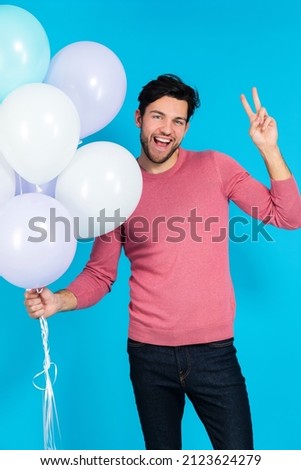 Smiling Funny Caucasian Guy Handsome Brunet Man With Bunch of Colorful Air Balloons Posing in Pink Jumper While Gesturing V-Signe Against Blue Background.Vertical Image