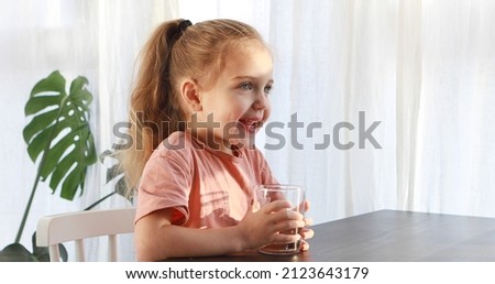 Little blonde girl in white dress drinks water from a glass indoors sunny day. Cute child is drinking a cup of water