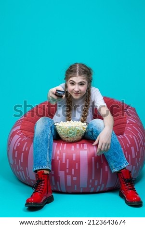 Attractive And Cheerful While Excited Teen Girl Sitting in Chair With TV Remote Control Changing Programs Online Isolated on Turquoise Trendy Vivid Vibrant Color Background.Vertical image