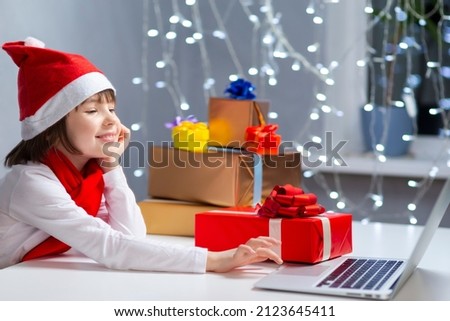 Smiling Caucasian Girl In Santa Hat Working With Laptop for Online Video Classroom Chat While Showing Wrapped Red Giftbox During Study From Home. Horizontal Image Composition