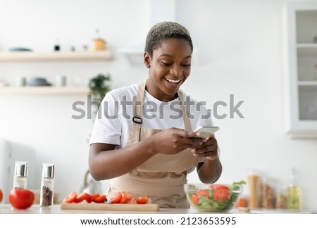 Smiling young pretty african american woman blogger in apron making photo of salad for food blog in kitchen interior, copy space. Video lesson, cooking meal, app for recipe during covid-19 lockdown Royalty-Free Stock Photo #2123653595