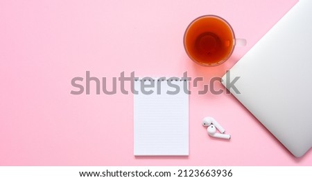 Notepad with a blank white page next to a silver closed laptop and a cup of tea on a pink background. Flat lay, copy space. Women's workplace.