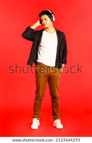 Close up shot of Asian handsome male model wearing headphones and posing for a photographing by bad mood isolated on red background.