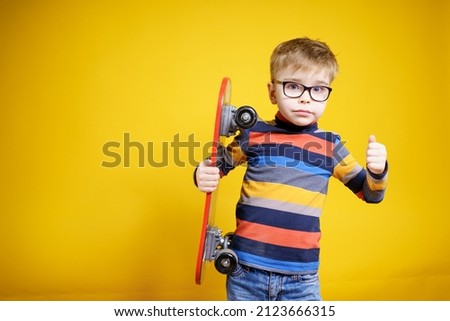 A boy with glasses in a sweater and jeans on a yellow background, holds a skateboard. Funny schoolboy. Hobby. Leisure.