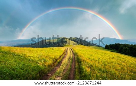 Amazing scene in summer mountains. Lush green grassy meadows in fantastic evening sunlight. Rural road and beautyful rainbow in dramatic sky. Landscape photography panorama Royalty-Free Stock Photo #2123671244