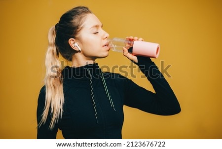Attractive caucasian girl on yellow isolated background with wireless headphones in sport suit make workout, run and drinking water