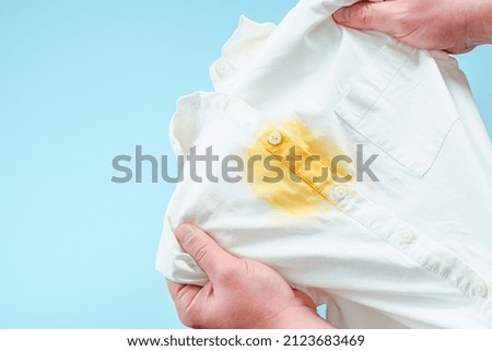 Dirty yellow stain on clothes. men's hands showing a white shirt. space for text. isolated. on blue background. top view. High quality photo Royalty-Free Stock Photo #2123683469