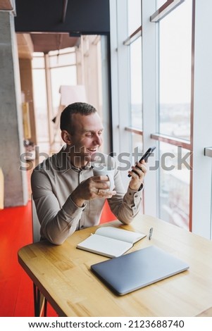 Successful young man is sitting at a table in a cafe-restaurant indoors, working and studying on a laptop computer,holding a mobile phone with a blank screen. Freelancer mobile office business concept