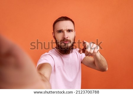 Portrait of serious handsome bearded man making selfie or streaming, pointing to camera, POV, point of view of photo, wearing pink T-shirt. Indoor studio shot isolated on orange background.