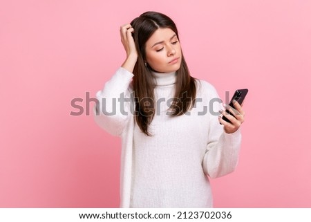 Pensive young woman holding smart phone in hands, having thoughtful facial expression, touches head, wearing white casual style sweater. Indoor studio shot isolated on pink background. Royalty-Free Stock Photo #2123702036