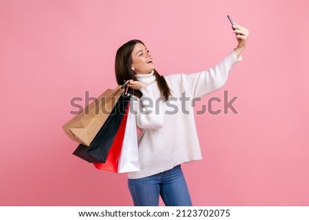 Happy excited dark haired female posing with shopping bags, making selfie, boasting her purchases, wearing white casual style sweater. Indoor studio shot isolated on pink background.