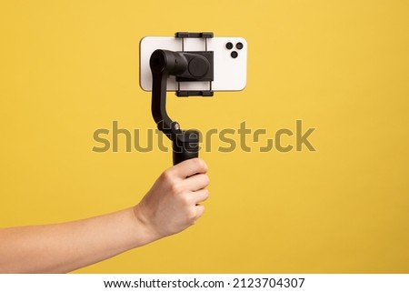 Closeup side view of woman hand holding steadicam with phone, for making video or has livestream. Indoor studio shot isolated on yellow background. Royalty-Free Stock Photo #2123704307