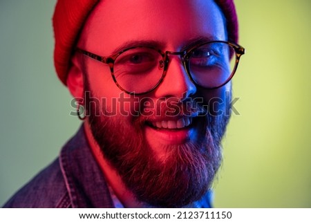 Closeup portrait of happy bearded hipster man looking at camera with toothy smile, optimism, success, wearing red beanie hat. Indoor studio shot isolated on colorful neon light background.