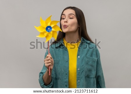 Hand mill. Portrait of happy beautiful young adult woman blowing at paper windmill, pinwheel toy on stick, wearing casual style jacket. Indoor studio shot isolated on gray background. Royalty-Free Stock Photo #2123711612