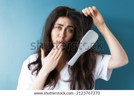 A confused young woman shows her long tangled hair, in which a hairbrush is stuck. Blue background. The concept of unruly hair care.