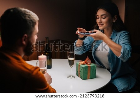 Back view to happy young woman taking picture on mobile phone of gift given by boyfriend in celebration of birthday or Valentine Day holiday. Cheerful couple husband and wife celebrate anniversary.
