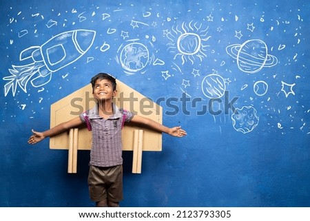 happy kid with cardboard rocket on back with space, universe and planets doodle drawing on wall - concept showing of childhood dream about astronaut or scientist Royalty-Free Stock Photo #2123793305