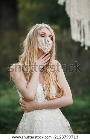 portrait of a bride in a mask posing. Beautiful young bride in a wedding dress in the park. On her face she has a fashionable white mask with pebbles