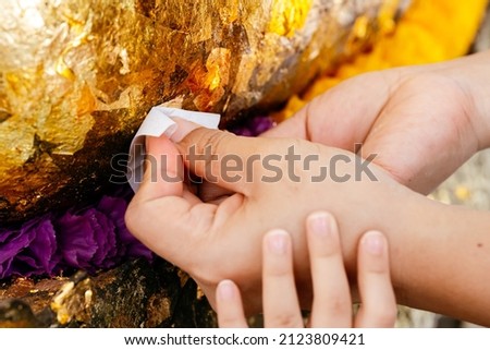 Hands mother and daughter affixing gold leaf to  Buddha statue. Kid girl hand touching onto hand adult holding gold leaf wrapped in paper. Making merit and paying respect to monks Buddhist beliefs. Royalty-Free Stock Photo #2123809421