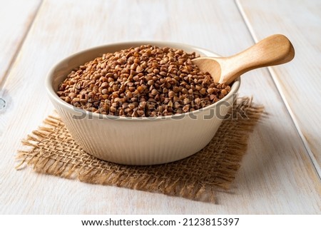 Beige ceramic bowl full of raw wholegrain buckwheat groats and spoon on a white wooden table. Cooking buckwheat porridge. Concepts of gluten free diet and vegetarian food. Close-up. Royalty-Free Stock Photo #2123815397