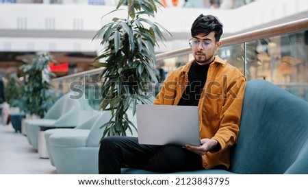 Young serious arab male freelancer in glasses sits looking at laptop screen works remotely loaded in thoughts about work thinks about business problem looks for inspiration new ideas finds solution Royalty-Free Stock Photo #2123843795