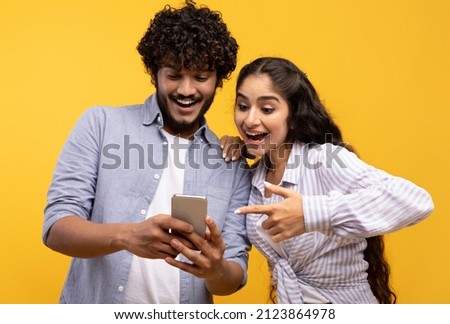 Portrait of excited indian couple using cellphone and pointing on it, looking at phone with open mouth, posing over yellow background, banner