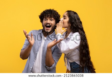 Young indian woman sharing secrets with her excited boyfriend, millennial couple whispering gossips to each other over yellow studio background Royalty-Free Stock Photo #2123865038