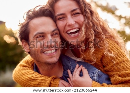 Portrait Of Happy Loving Couple With Man Giving Woman Piggyback As They Hug In Autumn Park Together