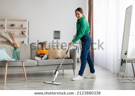Happy young mother mopping the floor and tidying up while her little son sitting on sofa and watching cartoon on smartphone, copy space