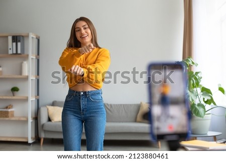 Young Caucasian female influencer recording video on smartphone, dancing on camera at home. Positive millennial lifestyle vlogger making content for her blog, filming for social media