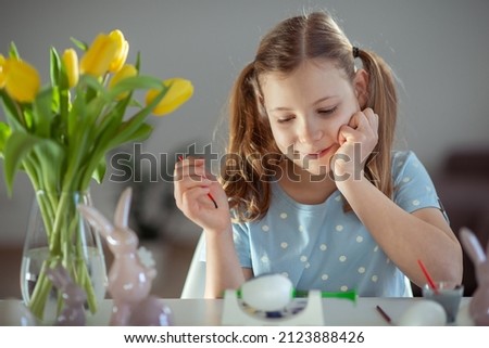 Adorable little girl with two funny pagtails smiling and painting easter eggs at table at home