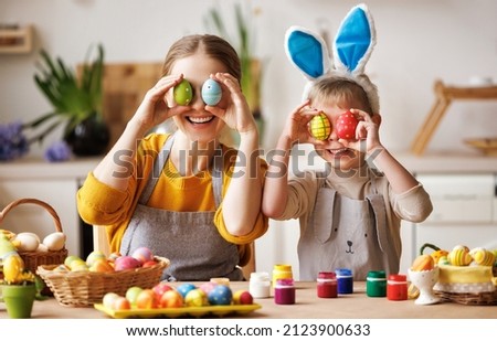 Happy mother and little son wearing aprons holding painted colorful Easter eggs in front of eyes while decorating them with food dyes in cozy kitchen at home. Easter craft activities for families Royalty-Free Stock Photo #2123900633