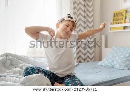Cute little kid boy wake up in the morning in his bed, stretching hand rise up to the air while sitting in sunny bedroom with big window on background. Royalty-Free Stock Photo #2123906711