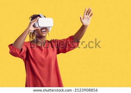 Happy excited Afro American woman in VR headset experiencing wonders of virtual reality. Black female gamer exploring new worlds in videogame. Studio shot, isolated on vibrant yellow mockup background