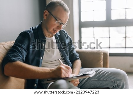 A man with glasses in casual clothes sitting on the couch writes something in a notebook